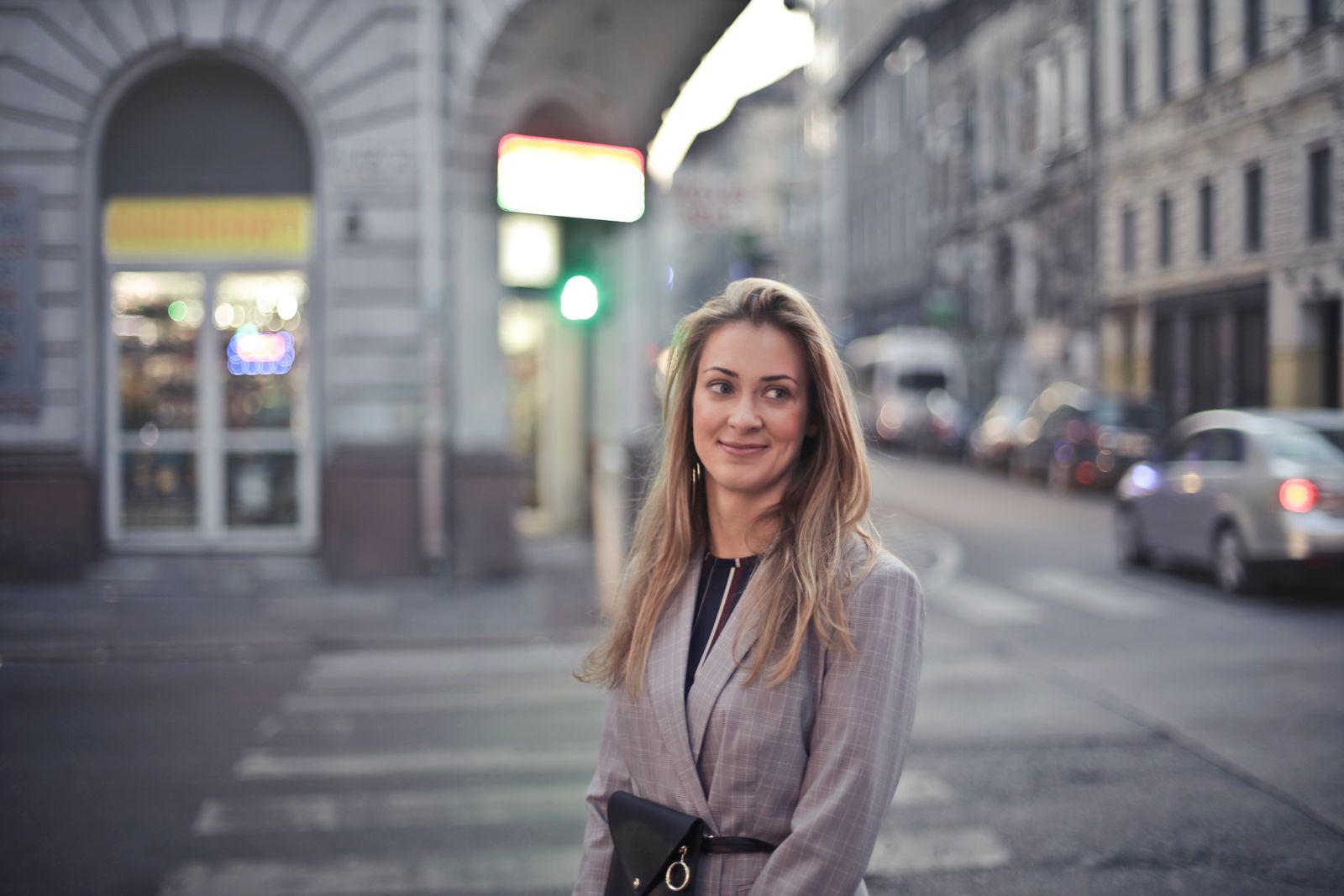 Eine lächelnde Frau im grauen Blazer steht auf einem Fussgängerstreifen in der Stadt, dahinter unscharfe Gebäude und ein Auto.
